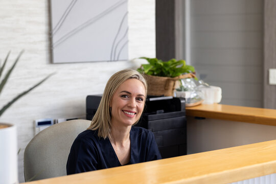 Mid-adult female receptionist sitting behind wood desk in modern office, with printer and plants