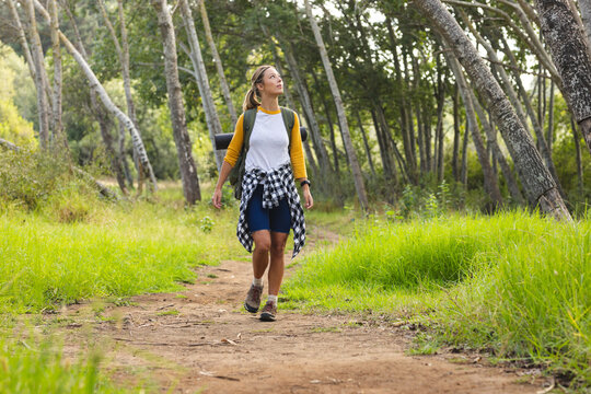 woman hiking on narrow forest trail under dappled sunlight, carrying green backpack and plaid shirt