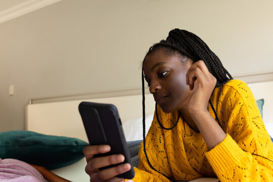 African American teenage girl lying on bed at home, holding smartphone with teal pillow, copy space