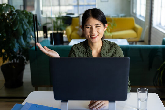 Asian woman sitting at desk in office lounge, gesturing and smiling during video calling on monitor