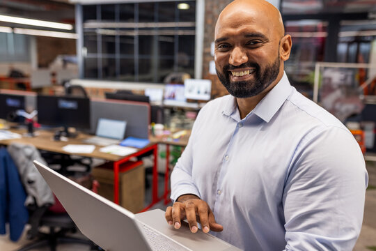 Mid-adult man using silver laptop in modern open-plan office with desks and monitors, copy space