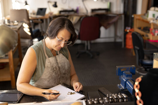 Senior woman sketching design in artisan workshop, using sketchbook pen with bench vise nearby