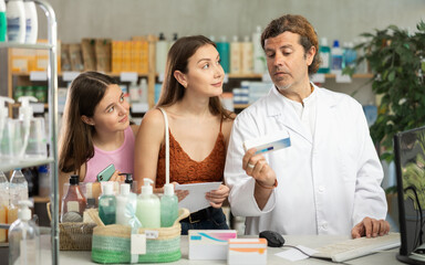 Man pharmacist using computer during patients consulting. Mother with teen child ask help with pick up medication according to doctors prescription in drug store, communicate with male staff. .