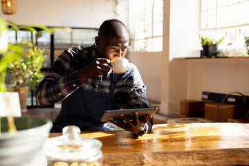 Young African American man sipping coffee and using tablet at sunlit cafe table with denim apron
