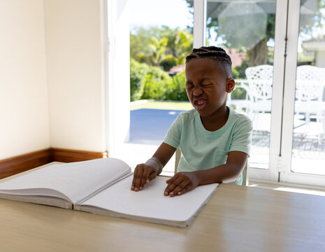 African American boy exploring Braille book at table in bright study room, tracing raised dots