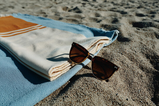 A single pair of modern sunglasses placed neatly on a folded beach towel with clean stripes, laid on light-colored sand