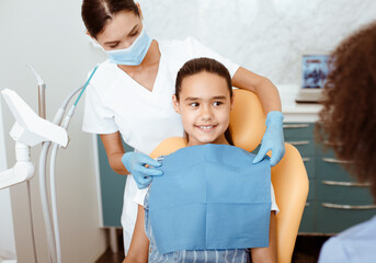 Fototapeta premium Service in modern dental clinic. Young female african american doctor looking at nurse in white coat, protective mask and gloves, dress up napkin on hispanic smiling small girl in chair, free space