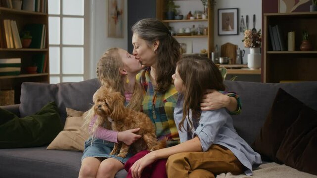 Medium shot of happy Caucasian single mom, teenage son and young daughter relaxing together on couch at family home with pet maltipoo dog, hugging, cuddling and kissing