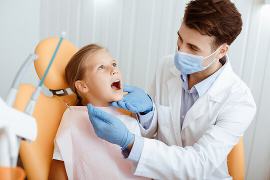 Dental care, routine check ups and pediatric dentistry. Millennial serious man in white coat, protective mask and gloves examines mouth of little kid in medical chair in modern clinic interior