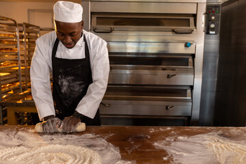 African American male baker rolling dough on floured countertop in bakery kitchen, with steel oven