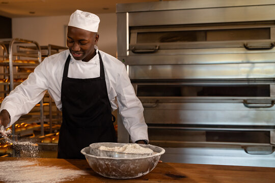 African American male baker sprinkling flour on countertop in bakery kitchen with metal mixing bowl