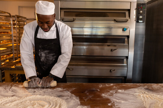 African American male baker rolling dough on floured countertop in bakery kitchen, with steel oven
