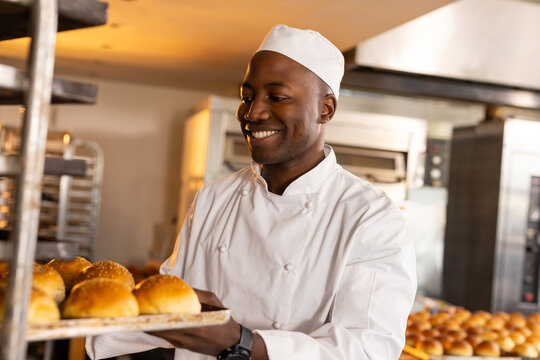 Young African American male chef pulling tray of fresh buns in bakery kitchen with ovens, smiling