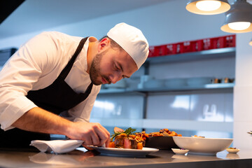 young male chef plating dish on stainless steel countertop in restaurant kitchen under lights