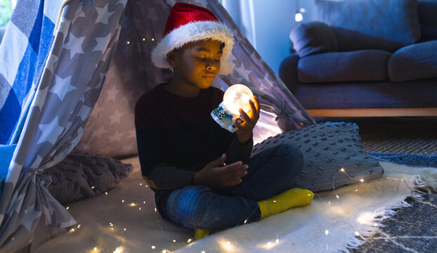 African American boy sitting in star teepee in living room with fairy lights, holding snow globe - Powered by Adobe