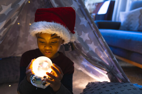 African American boy wearing Santa hat sitting in gray teepee in living room, holding glowing orb