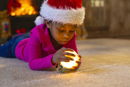 African American girl wearing Santa hat lying on carpet in living room, holding glowing orb