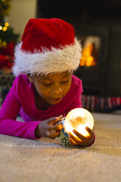 African American child lying in living room holding glowing Christmas orb, wearing Santa hat