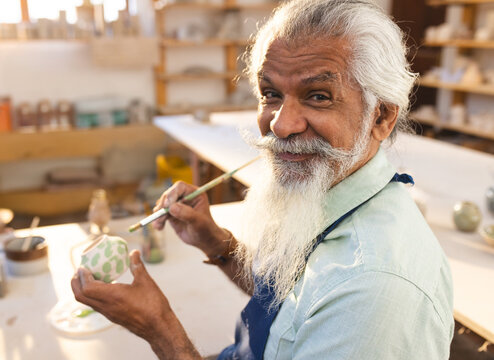 Senior man painting small ceramic pot at pottery studio, with fine paintbrush and glaze jars