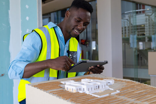 young African American man leaning over scale model in building site using tablet, copy space