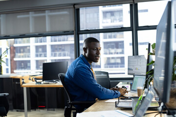 African American young man analyzing data on monitors in open-plan office, holding coffee cup