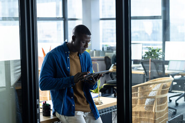 African American young adult man leaning on wooden desk in open-plan office, using tablet computer