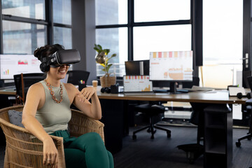 Asian woman using VR headset in wicker chair in office with monitors and laptop, copy space