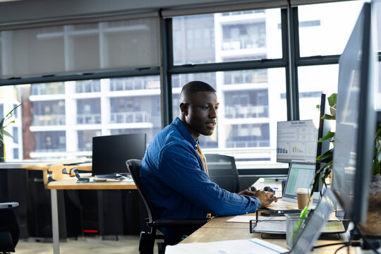 African American young man analyzing data on monitors in open-plan office, holding coffee cup
