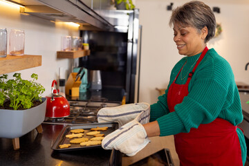 Woman removing cookie tray in home kitchen, with striped oven mitts and herb planter, copy space