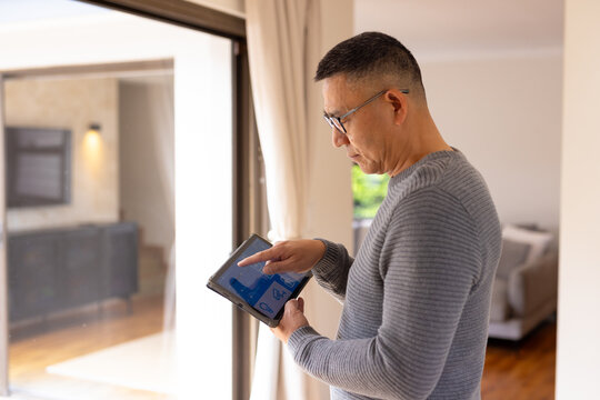 Senior Asian man tapping tablet in living area by sliding glass door, with charts, copy space - Powered by Adobe