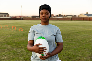 Teenage African American girl practicing rugby on field holding white green ball, wearing scrum cap