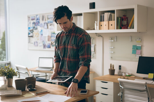 Young man reviewing tablet in home office, holding papers amid scattered desk items