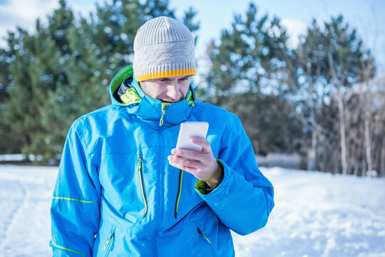 Woman standing in snowy clearing, holding white smartphone, wearing blue winter jacket and beanie