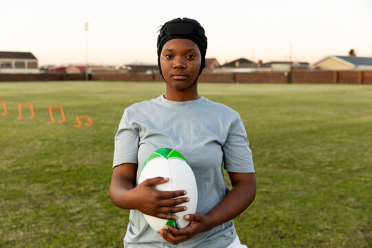 Teenage African American girl practicing rugby on field holding white green ball, wearing scrum cap