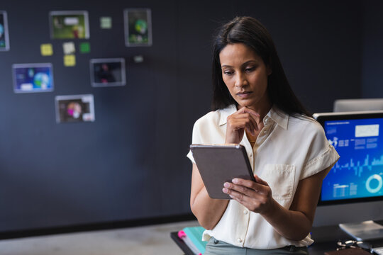 Woman holding tablet reviewing data charts on monitor in office with inspiration board, copy space