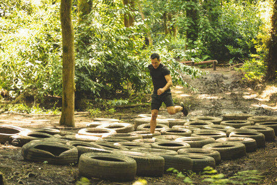 Man navigating rows of tires on muddy forest obstacle course, stepping over branches