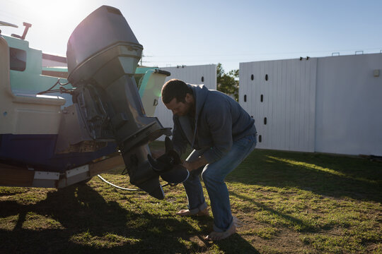 Man bending over outboard motor on trailer in backyard by white fence, adjusting motor, copy space