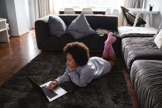 Woman lying on dark shaggy rug in living room, using silver laptop beside gray sectional sofa