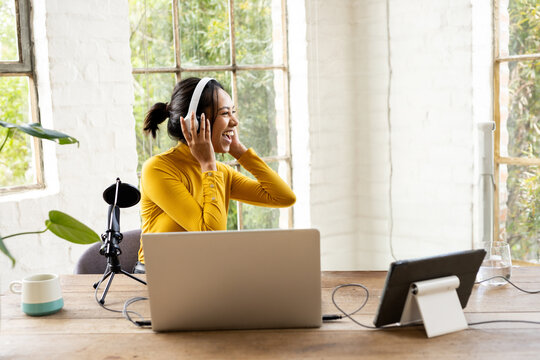 Woman adjusting white headphones and speaking into microphone at wooden desk in studio with laptop
