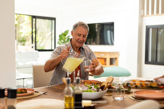 Senior man pouring lemonade from pitcher into glass at dining room table, with salad and utensils