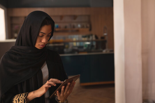 Young adult woman standing in modern café kitchen, tapping tablet while wearing wristwatch - Powered by Adobe