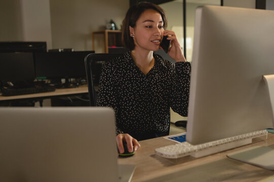 Young adult woman talking on smartphone at office desk with desktop monitor and wireless mouse