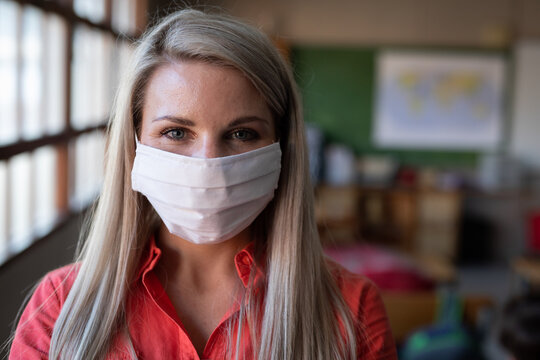 Female teacher standing facing camera in brightly lit classroom, with white mask, red blouse, map