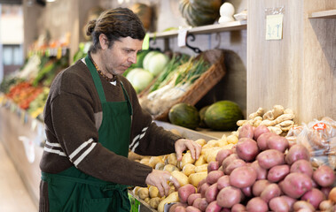 Adult male seller in uniform lays out potatoes on counter in vegetable store