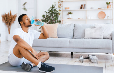 Stay Hydrated. Black Man Drinking Water During Workout, Exercising Sitting On Gymnastics Mat...