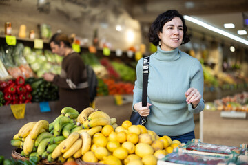 Middle-aged woman stands in a supermarket near a fruit stall and looks at the products with interest. Customer chooses fresh fruit in the grocery department