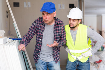 Focused creative adult male interior designer giving instructions to construction team foreman and making notes on drywall in building under renovation