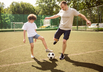 Happy Childhood Concept. Mature man enjoying soccer game with little boy on sports field at sunny day