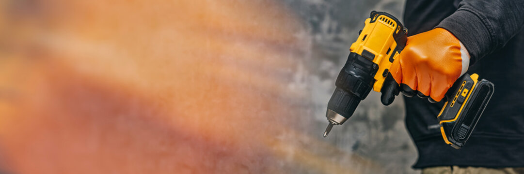 A construction worker in orange gloves holds a power drill against a concrete wall. Concept construction and renovation themes.