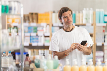 Fototapeta premium Adult man with mobile phone choosing medicines and hygiene products in pharmacy